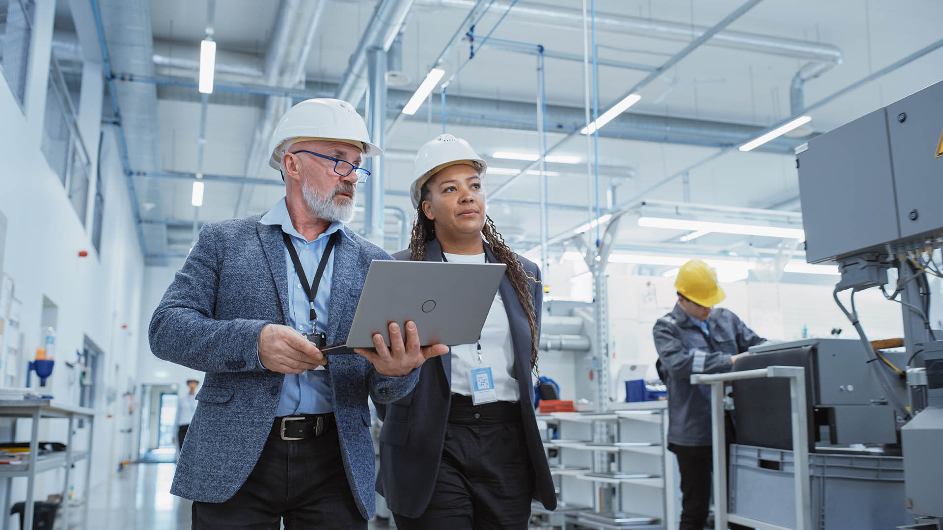 Two Professional Heavy Industry Engineers Wearing Hard Hats at Factory. Walking and Discussing Industrial Machine Facility, Working on Laptop. African American Manager and Technician at Work.